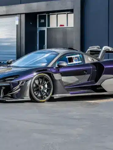 Seitliche Ansicht eines McLaren Senna GTR in dunklem Violett mit markantem Heckflügel und Rennsportdetails, geparkt vor einem modernen Industriegebäude.