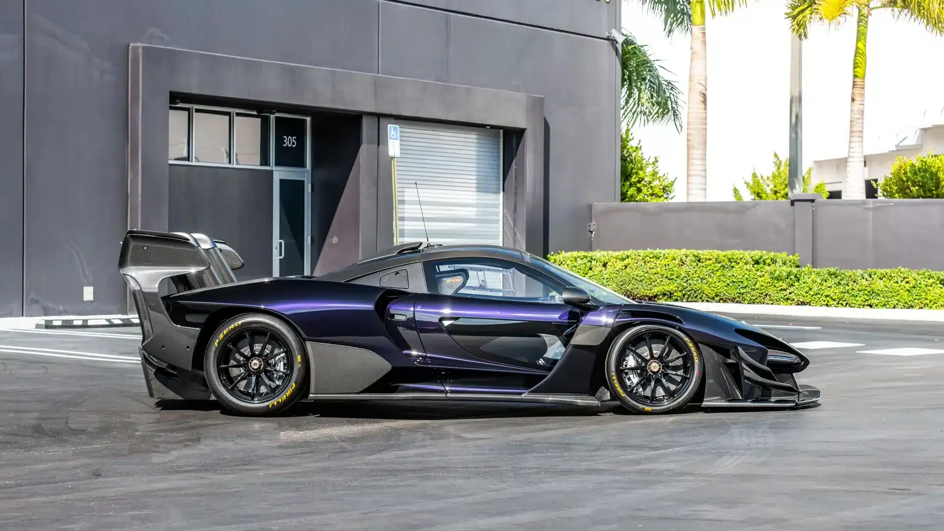 Seitenansicht eines McLaren Senna GTR in dunkler Lackierung, mit markantem Heckflügel und Rennslicks, geparkt vor einem modernen Gebäude.