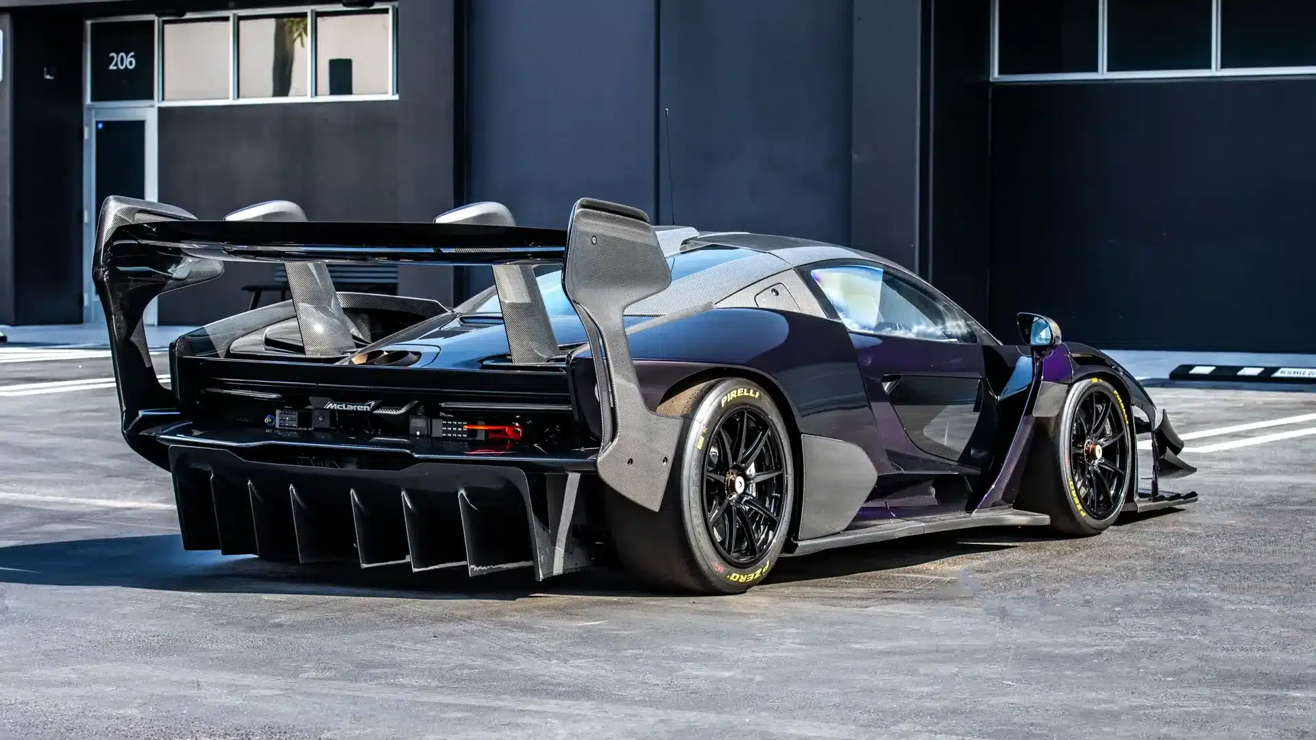 Heckansicht eines McLaren Senna GTR mit großem Heckflügel und Pirelli-Rennreifen, geparkt vor einem modernen Gebäude.