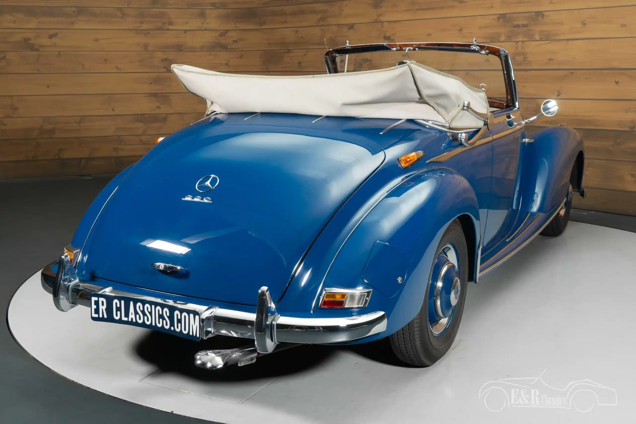 Heckansicht eines blauen Mercedes-Benz 220 A Cabriolet von 1951 mit geöffnetem, beigefarbenem Verdeck vor einer Holzvertäfelung.