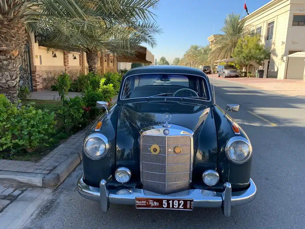 Frontansicht eines Mercedes-Benz 220 S Ponton, geparkt auf einer Straße mit Palmen und Häusern im Hintergrund.