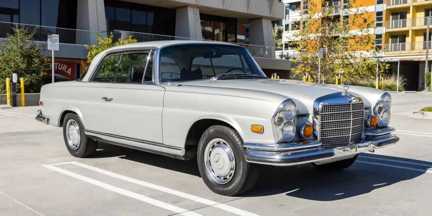 Silbergraues Mercedes-Benz 280SE Coupé W111 von 1970, geparkt im Freien vor einem modernen Gebäude, Seiten- und Frontansicht.