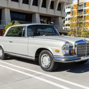 Silbergraues Mercedes-Benz 280SE Coupé W111 von 1970, geparkt im Freien vor einem modernen Gebäude, Seiten- und Frontansicht.