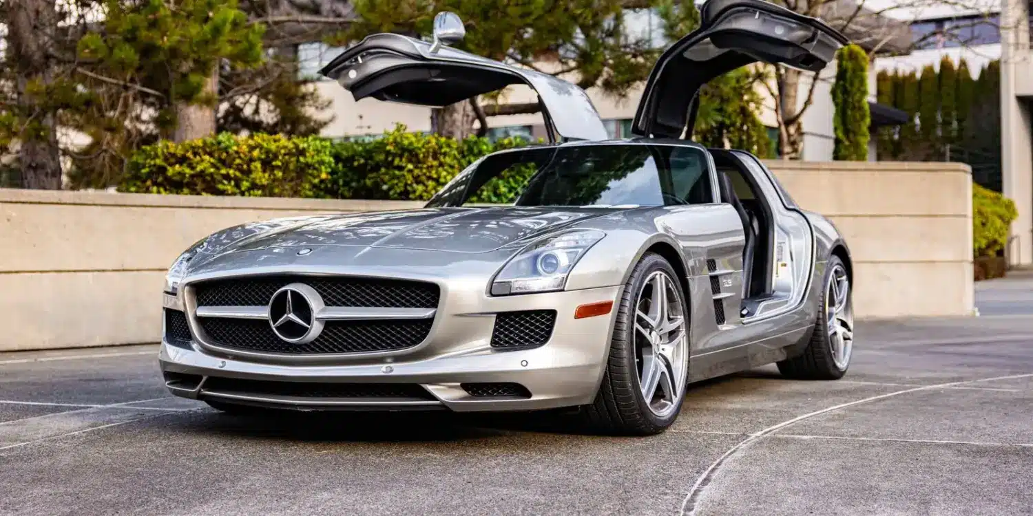 Silberner Mercedes-Benz SLS AMG mit geöffneten Flügeltüren auf einer Einfahrt, seitliche Frontansicht