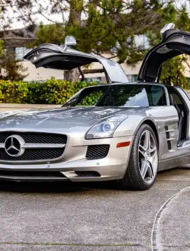 Silberner Mercedes-Benz SLS AMG mit geöffneten Flügeltüren auf einer Einfahrt, seitliche Frontansicht