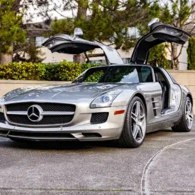 Silberner Mercedes-Benz SLS AMG mit geöffneten Flügeltüren auf einer Einfahrt, seitliche Frontansicht