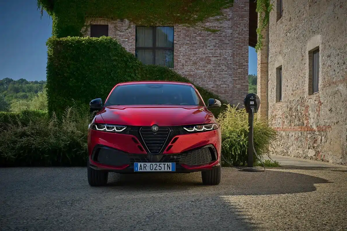 Frontansicht eines roten Alfa Romeo Tonale vor einem Gebäude mit Ladestation.