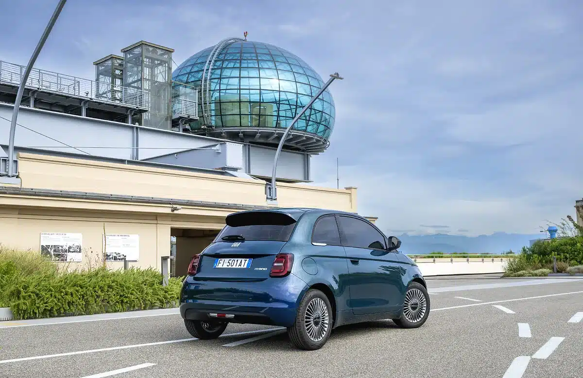 Fiat 500 Hybrid in Blau, fotografiert vor dem Mirafiori-Werk in Turin mit moderner Glaskuppel im Hintergrund.
