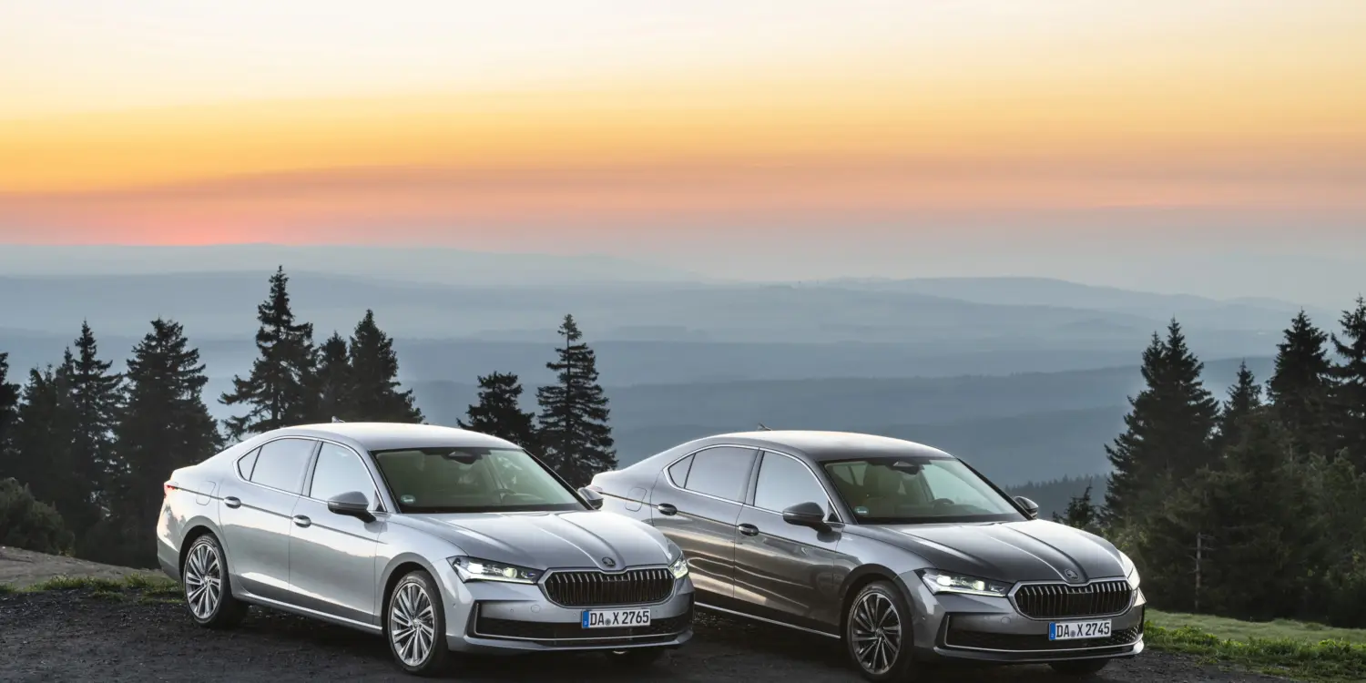 Zwei Škoda Superb 2025, Limousine und Combi, stehen nebeneinander bei Sonnenuntergang auf einem Hügel mit Blick auf bewaldete Täler.