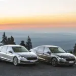 Zwei Škoda Superb 2025, Limousine und Combi, stehen nebeneinander bei Sonnenuntergang auf einem Hügel mit Blick auf bewaldete Täler.
