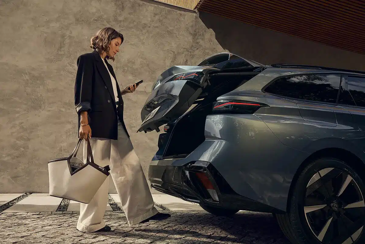 Frau steht neben einem silbernen Peugeot 308 SW mit geöffnetem, elektrisch betätigtem Kofferraumdeckel.