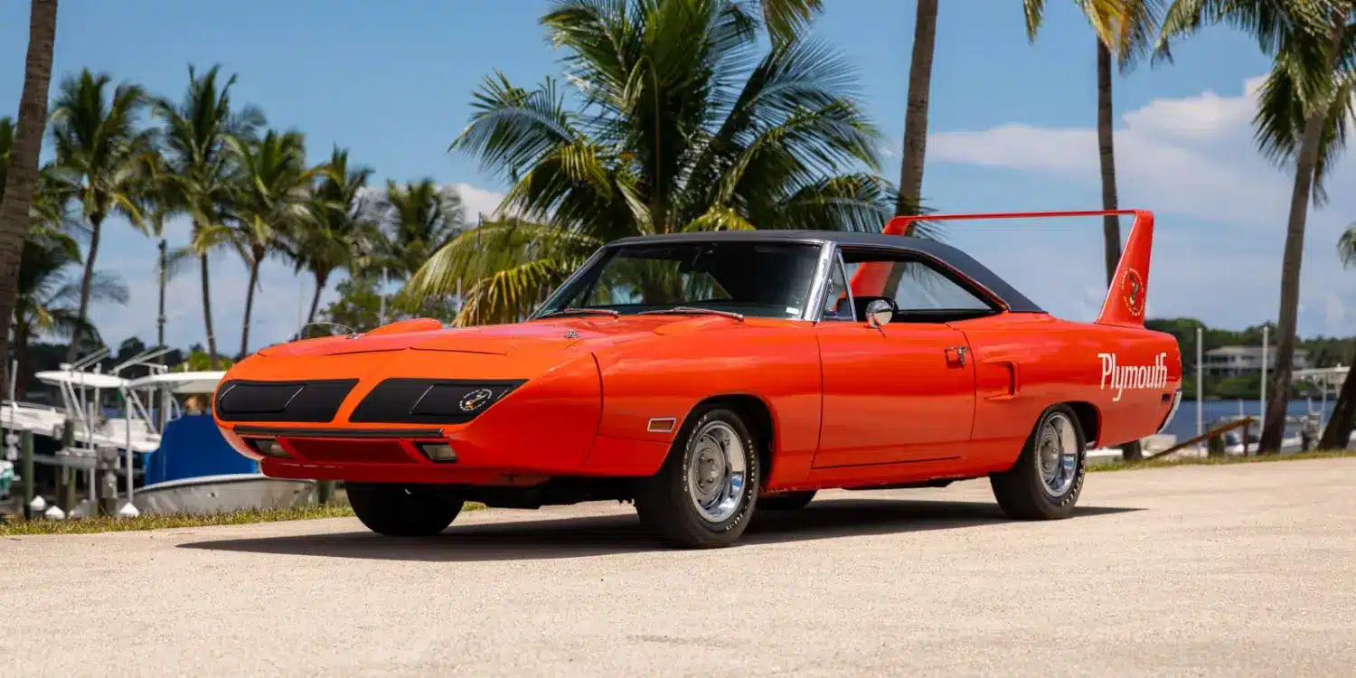 Leuchtend orangefarbener 1970 Plymouth Superbird mit charakteristischem, hohem Heckflügel und schwarzem Dach vor tropischer Kulisse.
