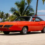 Leuchtend orangefarbener 1970 Plymouth Superbird mit charakteristischem, hohem Heckflügel und schwarzem Dach vor tropischer Kulisse.