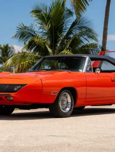 Leuchtend orangefarbener 1970 Plymouth Superbird mit charakteristischem, hohem Heckflügel und schwarzem Dach vor tropischer Kulisse.