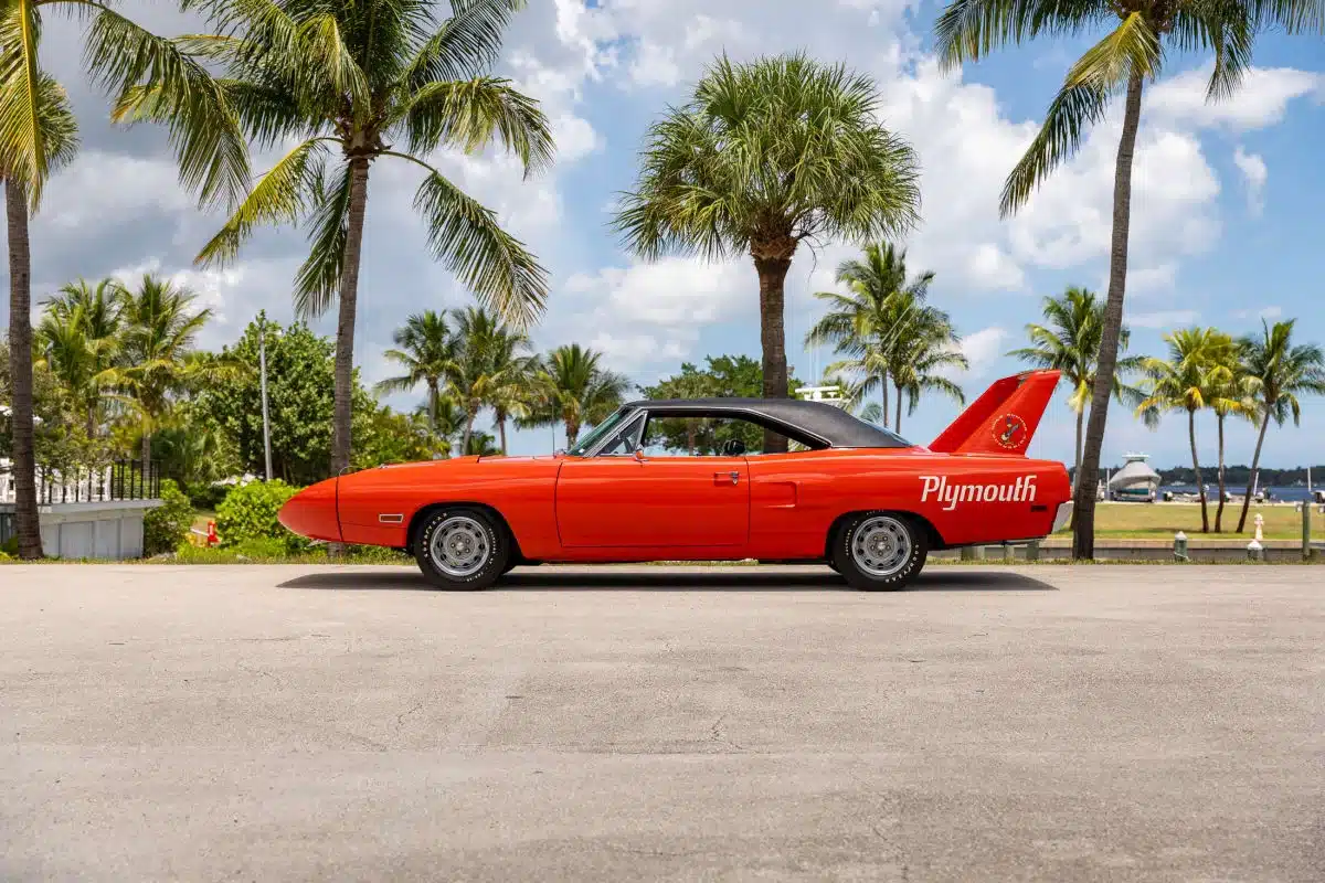 Seitliche Ansicht eines orangefarbenen 1970 Plymouth Superbird mit großem Heckflügel unter Palmen.
