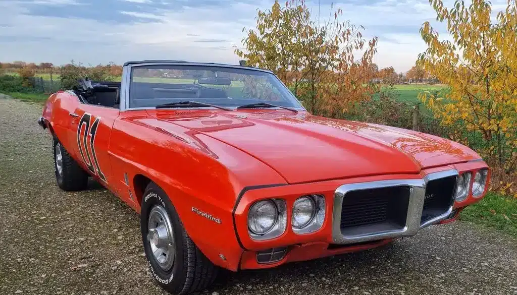 Pontiac Firebird Cabriolet 1969, orange lackiert, mit geöffnetem Verdeck und charakteristischer Front, stehend auf einem ländlichen Weg.