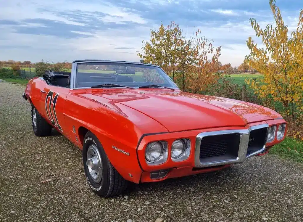 Pontiac Firebird Cabriolet 1969, orange lackiert, mit geöffnetem Verdeck und charakteristischer Front, stehend auf einem ländlichen Weg.