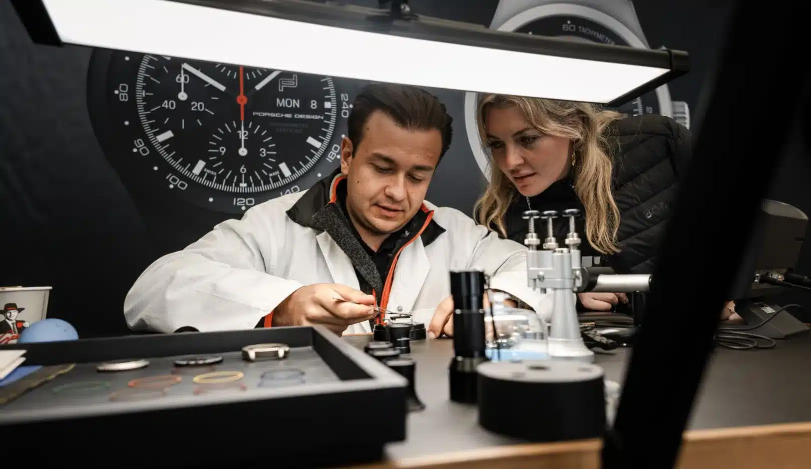 Zwei Personen bei der Montage eines Porsche Design Chronographen unter einer Werkbankleuchte, mit großem Ziffernblatt im Hintergrund
