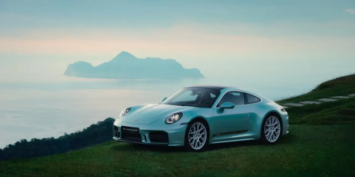 Porsche 911 Carrera T Formosa in Ipanema Blue Metallic auf einer grünen Wiese mit Küste und Meer im Hintergrund.