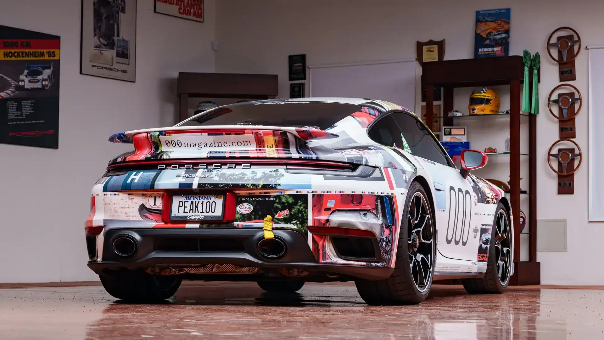 Individuell folierter Porsche 911 Turbo S Lightweight 2022 von hinten in einer Garage mit Porsche-Postern und Trophäen im Hintergrund.