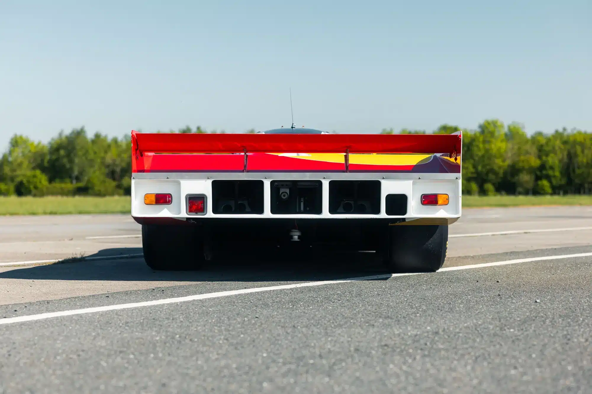Heckansicht des Porsche 962-010 mit großem Heckflügel und Shell/Dunlop-Rennlackierung auf einer Straße.