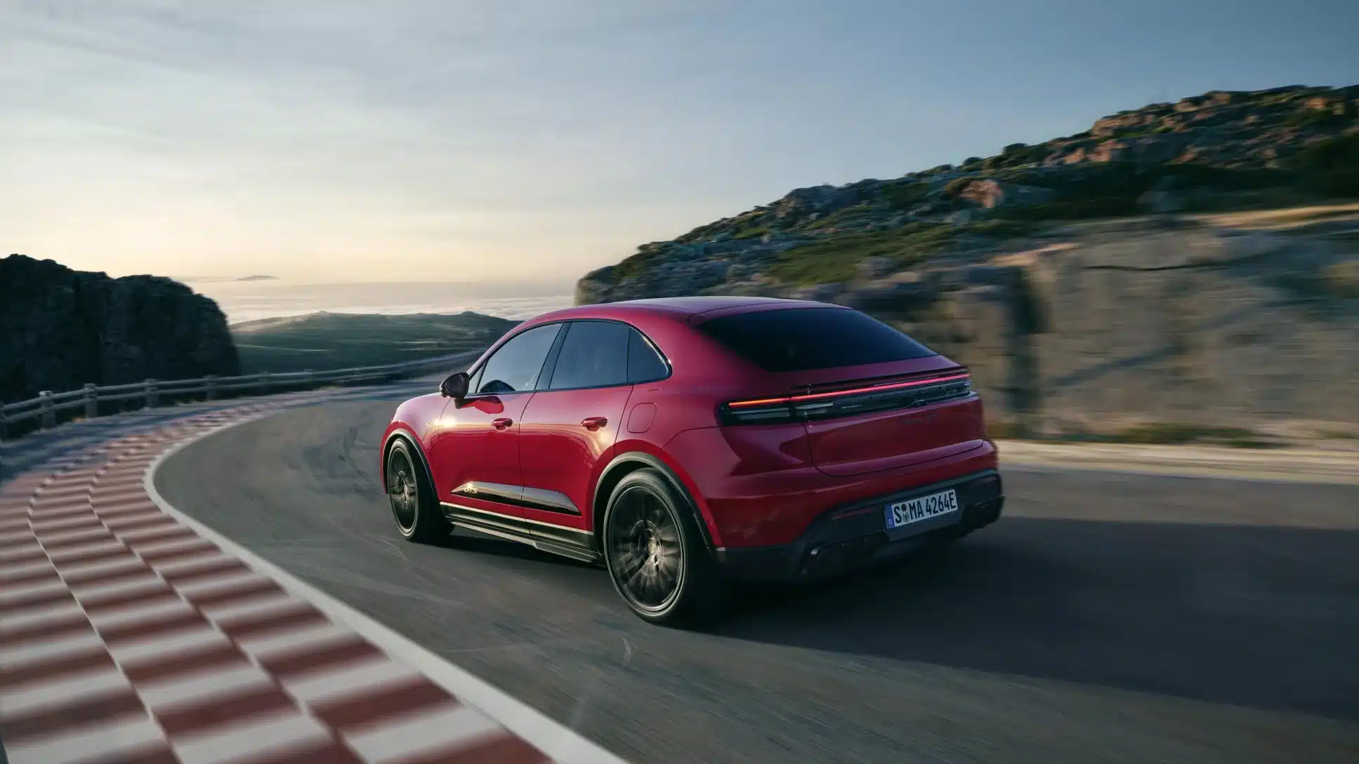 Roter Porsche Macan GTS Electric fährt dynamisch auf einer kurvigen Straße in bergiger Landschaft.