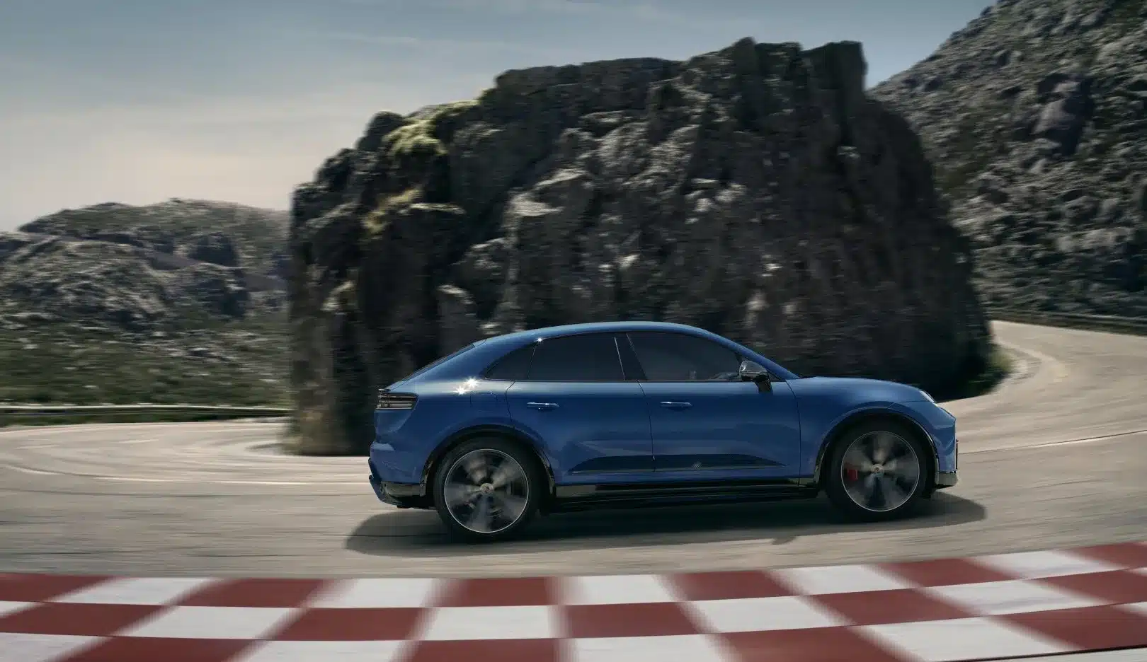 Blauer Porsche Macan GTS Electric fährt dynamisch auf einer Bergstraße mit Felsen im Hintergrund.