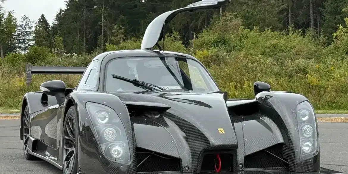 Frontansicht eines Radical RXC TwinTurbo GT 650R mit geöffneter Fahrertür, Kohlefaser-Karosserie, auf einer Straße vor bewaldetem Hintergrund