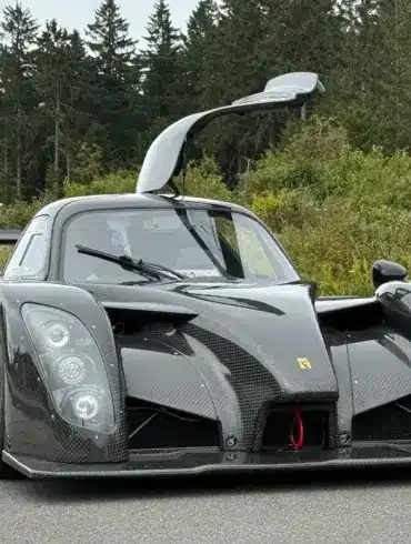 Frontansicht eines Radical RXC TwinTurbo GT 650R mit geöffneter Fahrertür, Kohlefaser-Karosserie, auf einer Straße vor bewaldetem Hintergrund