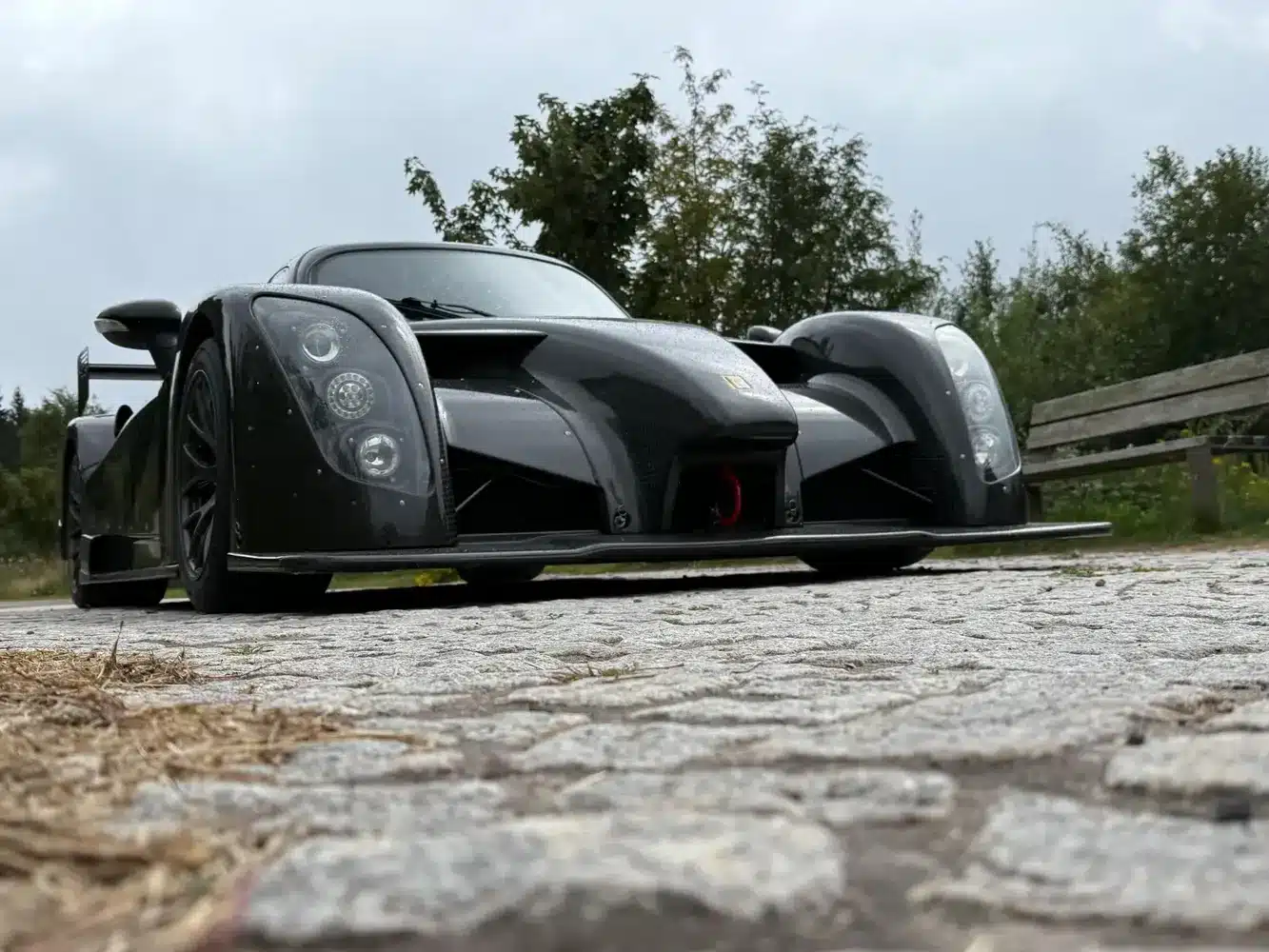 Radical RXC TwinTurbo GT 650R, schwarzes zweisitziges Coupé mit aerodynamischem Design, von vorne fotografiert auf Kopfsteinpflaster mit Hintergrund aus Bäumen und Bank.
