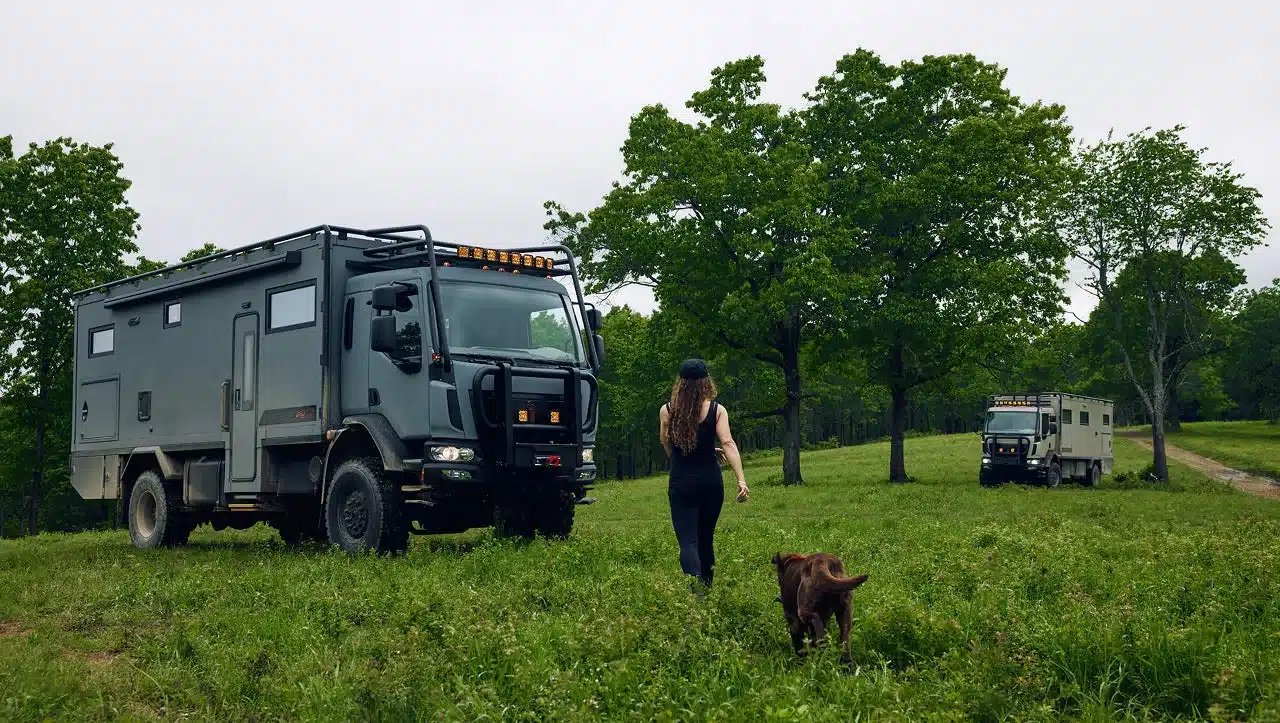 Ein Storyteller Overland GXV EPIC Expeditionsfahrzeug, das auf einer grünen Wiese steht. Im Vordergrund läuft eine Frau mit Hund auf das Fahrzeug zu.