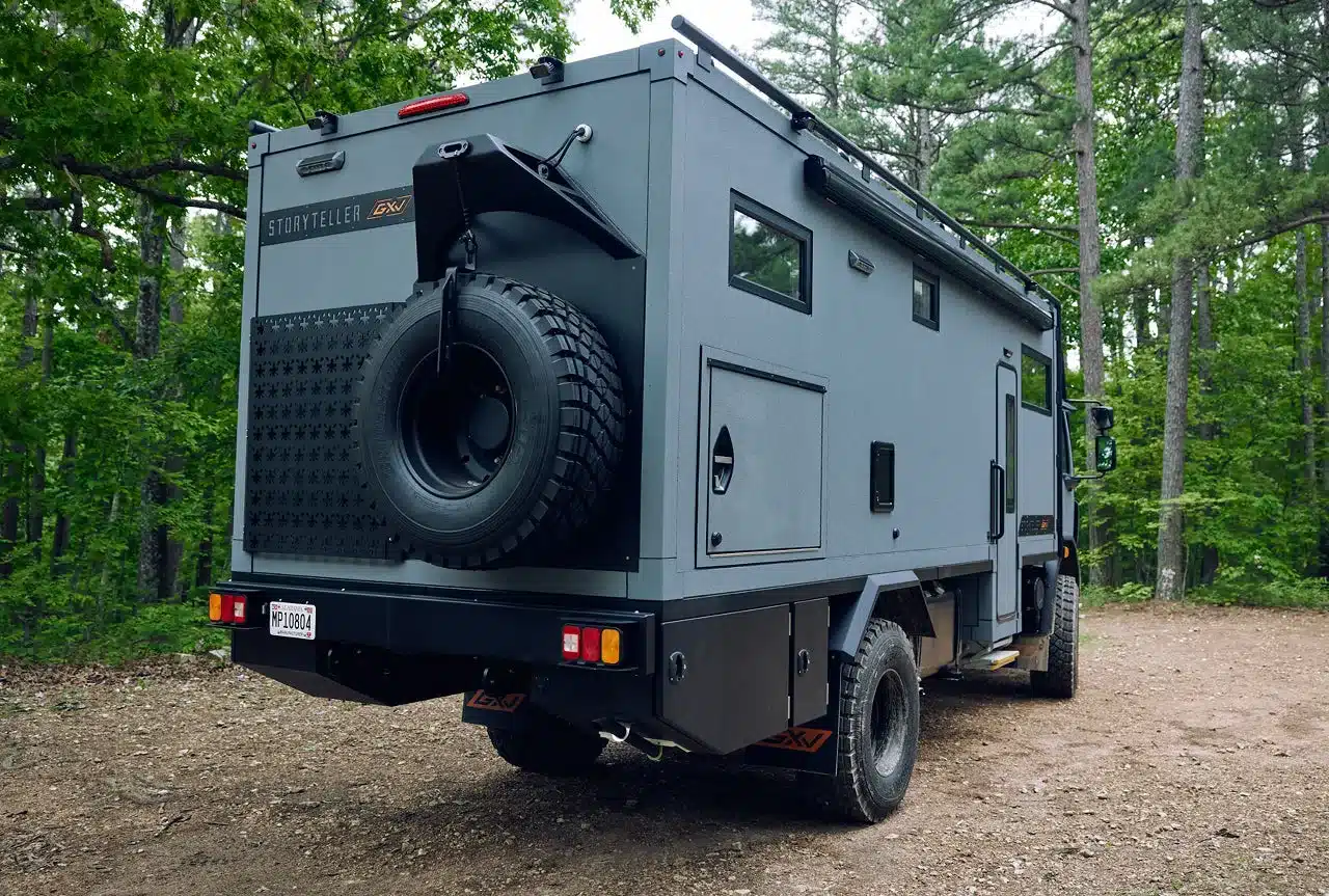 Storyteller Overland GXV EPIC von hinten im Wald, Ersatzrad am Heck, robuste Expeditionsausstattung sichtbar.