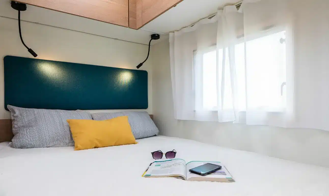 Schlafbereich mit festem Bett im Wohnmobil 60F, Tageslicht durch Seitenfenster, moderne Leselampen und Kissen