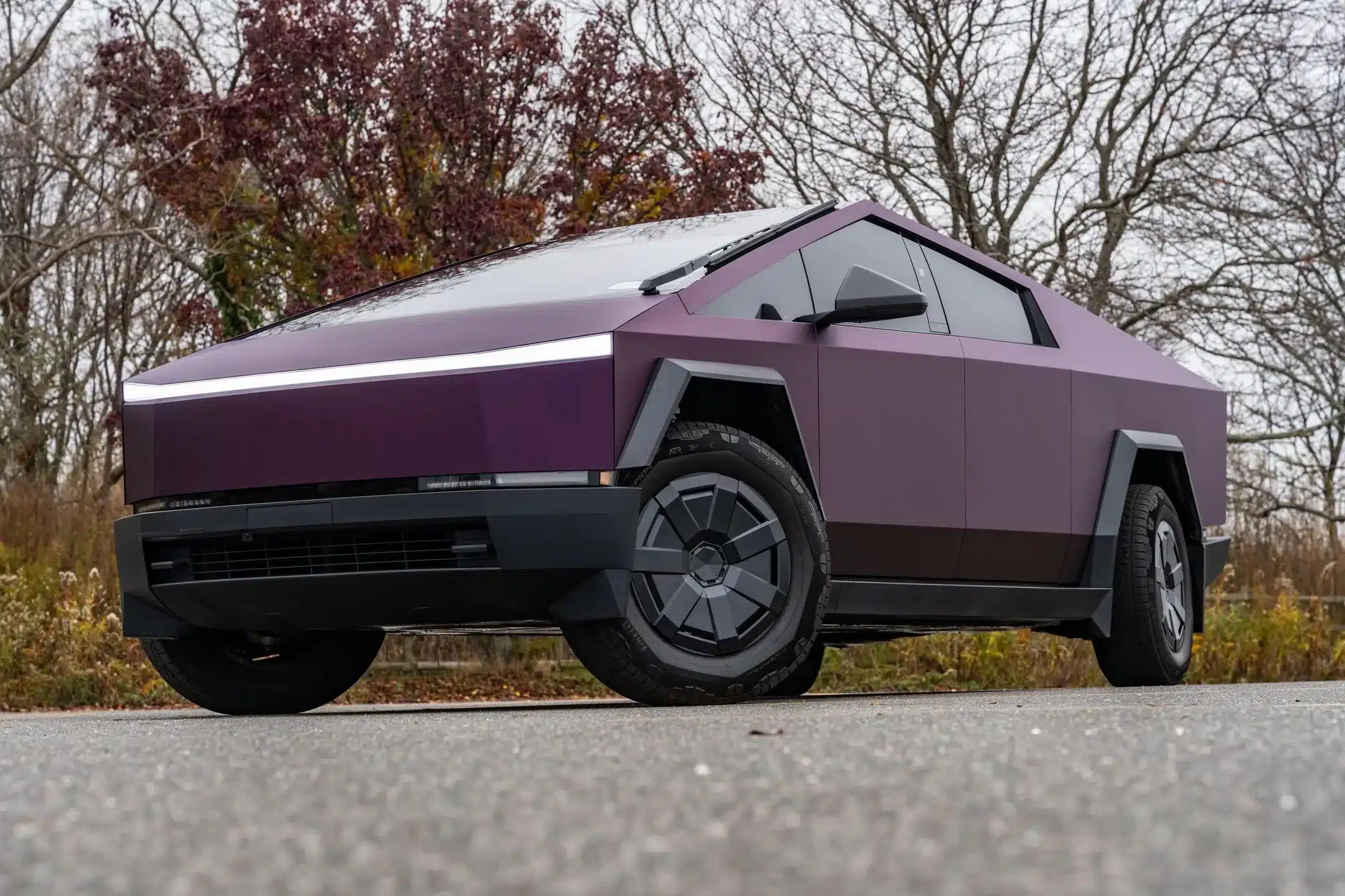 Tesla Cybertruck 2024 in violetter Folierung, fotografiert seitlich auf einer Straße mit Herbstbäumen im Hintergrund.