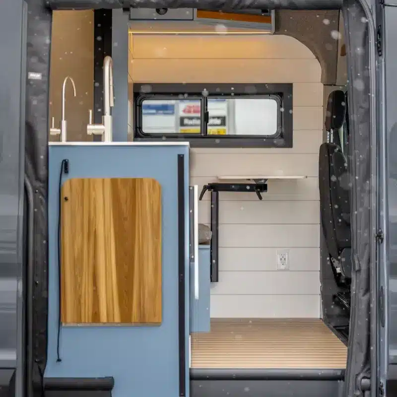 Innenraum eines Vancini Quetico Campervans mit Küchenzeile, Spüle, Holzoberflächen und flexiblem Tisch.