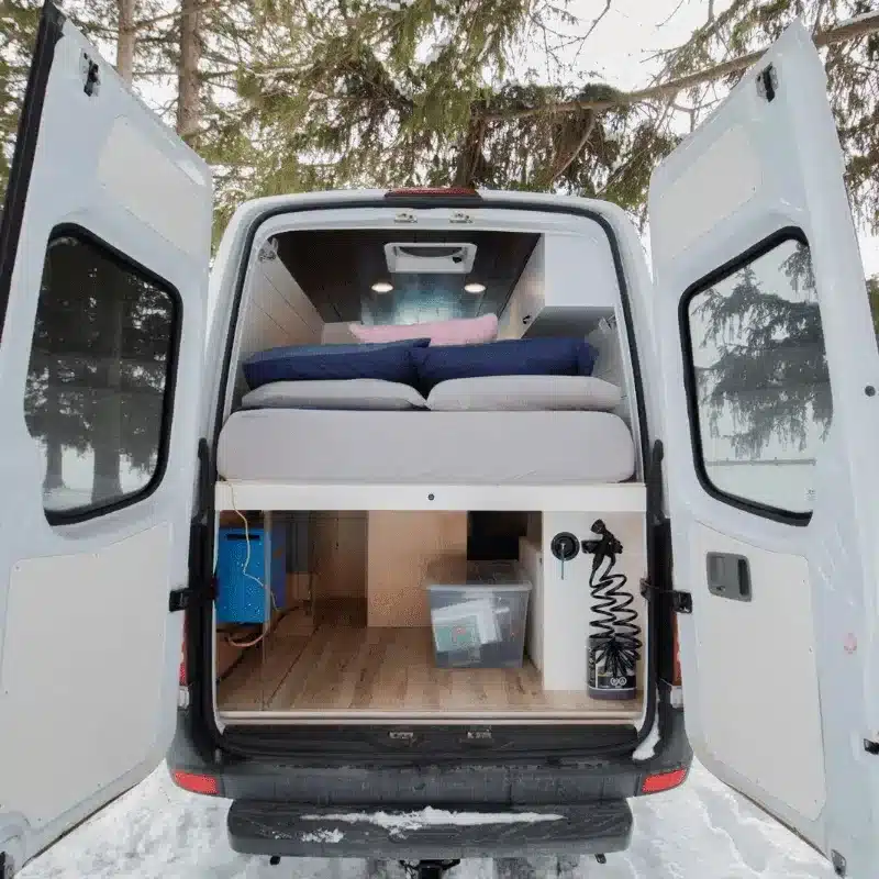 Heckansicht eines weißen Mercedes Sprinter Campers mit geöffneten Türen, herausnehmbarem Bett, Stauraum und Außendusche im Schnee.