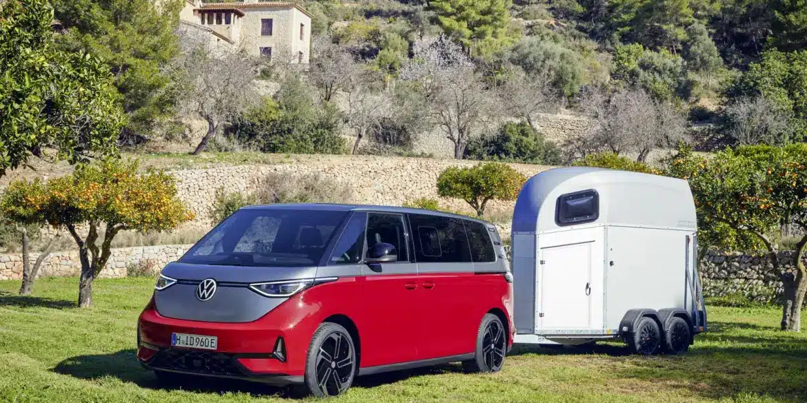Volkswagen ID. Buzz GTX in Kirschrot mit Anhänger auf einer Wiese, im Hintergrund mediterrane Landschaft und Haus.