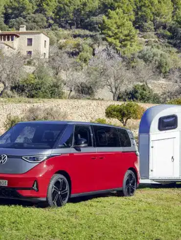Volkswagen ID. Buzz GTX in Kirschrot mit Anhänger auf einer Wiese, im Hintergrund mediterrane Landschaft und Haus.