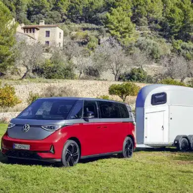 Volkswagen ID. Buzz GTX in Kirschrot mit Anhänger auf einer Wiese, im Hintergrund mediterrane Landschaft und Haus.