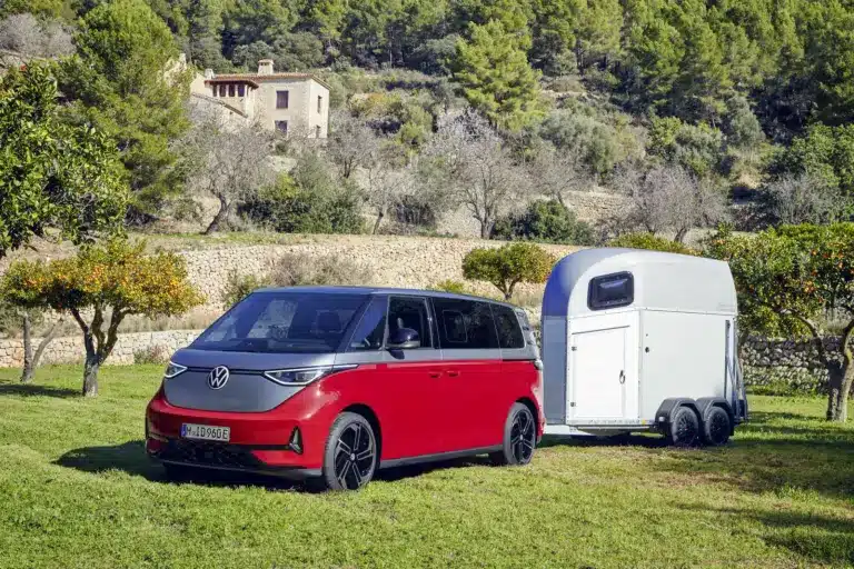 Volkswagen ID. Buzz GTX in Kirschrot mit Anhänger auf einer Wiese, im Hintergrund mediterrane Landschaft und Haus.