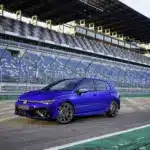 Blauer Volkswagen Golf R, frontal seitlich fotografiert, auf einer Rennstrecke mit Zuschauertribünen im Hintergrund.
