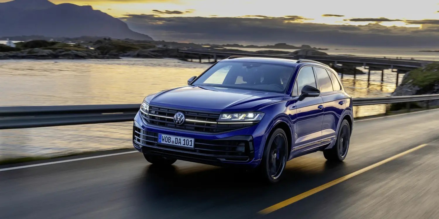 Blauer Volkswagen Touareg Final Edition fährt auf einer Straße vor malerischer Landschaft mit Wasser und Bergen im Sonnenuntergang.