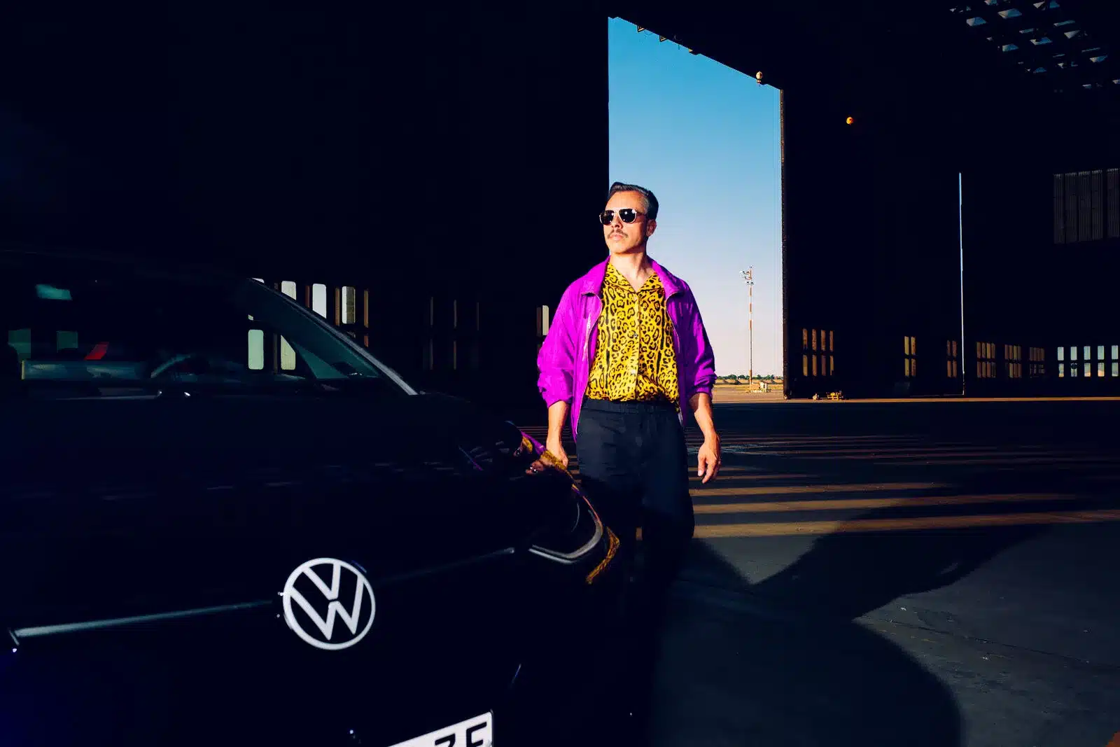 Ein Mann mit Sonnenbrille und auffälligem Retro-Outfit steht neben einem Volkswagen ID.3 GTX FIRE+ICE vor einem Hangar.
