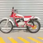 Seitenansicht einer Yamaha TY80 Trialsmaschine, rot-weiß lackiert, vor grauer Garagentor-Kulisse.