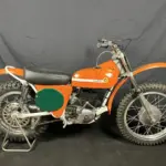 Seitenansicht einer restaurierten orangefarbenen Montesa 250 GP (Cappra) von 1968 vor dunklem Hintergrund.