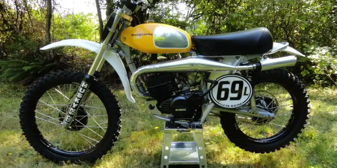 Seitenansicht einer Husqvarna 125CR Lightweight 1974 mit gelbem Tank, im Gelände auf einem Ständer platziert.