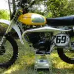 Seitenansicht einer Husqvarna 125CR Lightweight 1974 mit gelbem Tank, im Gelände auf einem Ständer platziert.