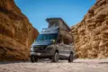 Mercedes Sprinter Kastenwagen PELE von Outpost Vans mit geöffnetem Aufstelldach und Offroad-Umbau, geparkt in einer felsigen Wüste.