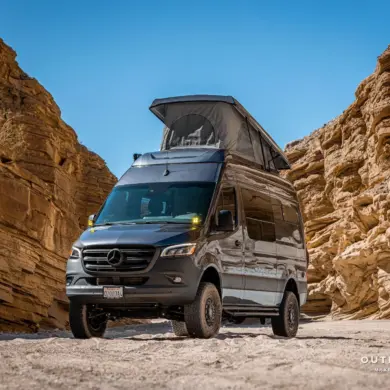 Mercedes Sprinter Kastenwagen PELE von Outpost Vans mit geöffnetem Aufstelldach und Offroad-Umbau, geparkt in einer felsigen Wüste.