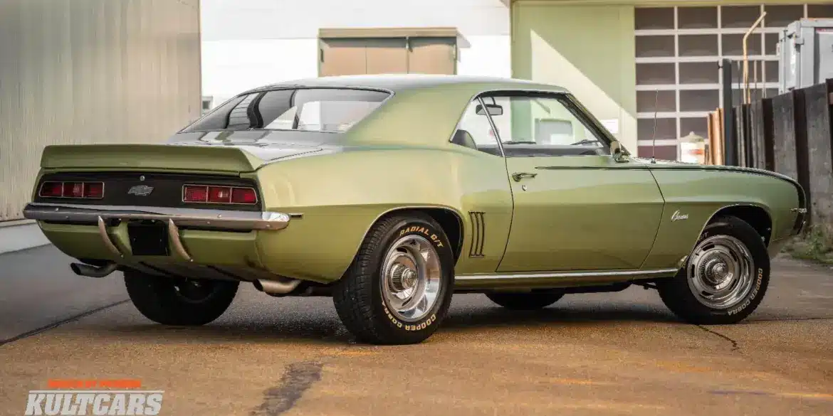 Seiten- und Heckansicht eines klassischen Chevrolet Camaro 350 V8 Coupé von 1969 mit grüner Metallic-Lackierung, geparktem Fahrzeug vor moderner Halle
